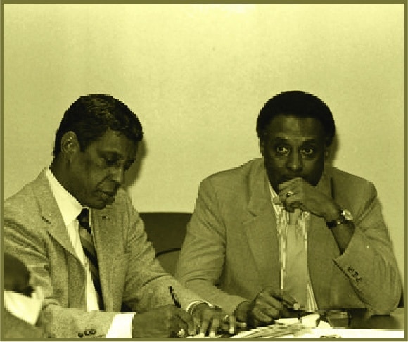 Sepia photo of two men in suits seated together, in conversation