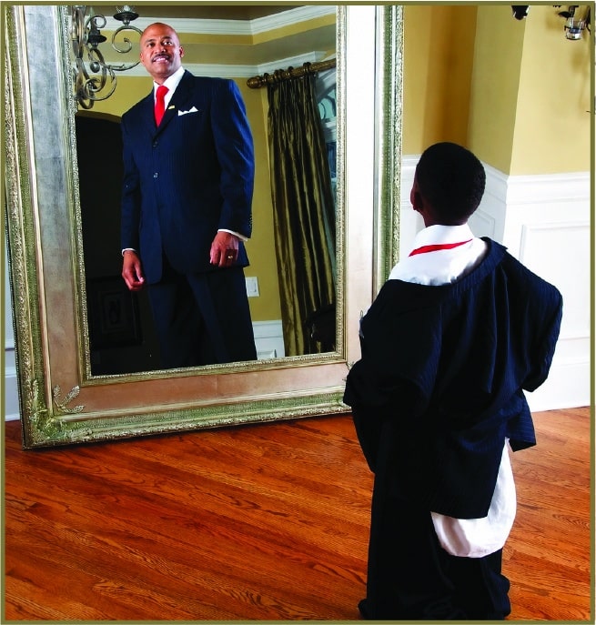 Photo of a young man in a graduation cap and gown looking at his reflection in a mirror